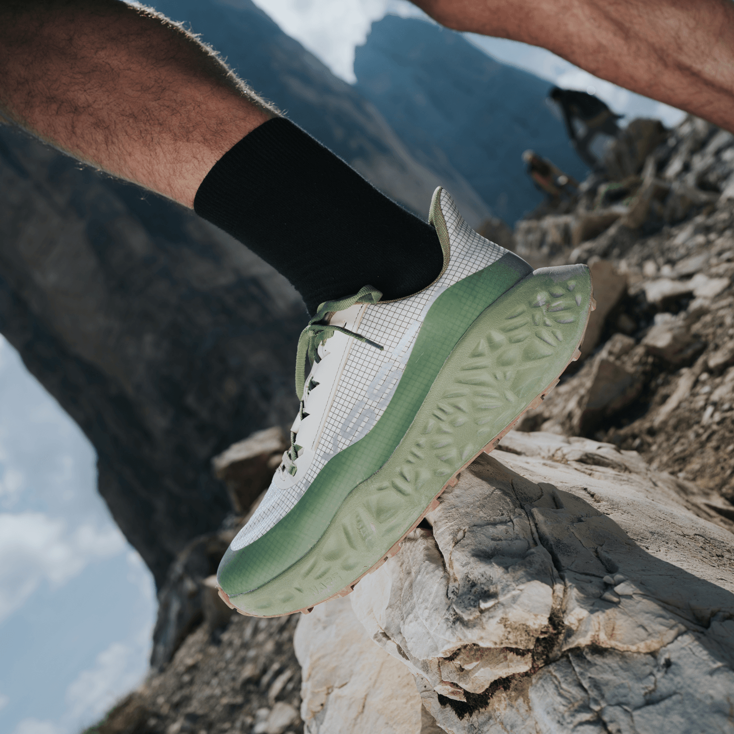 Close-up of a green and white Ulysses trail shoe with textured foam midsole, worn on a rocky mountain ridge with hikers visible in the distance.