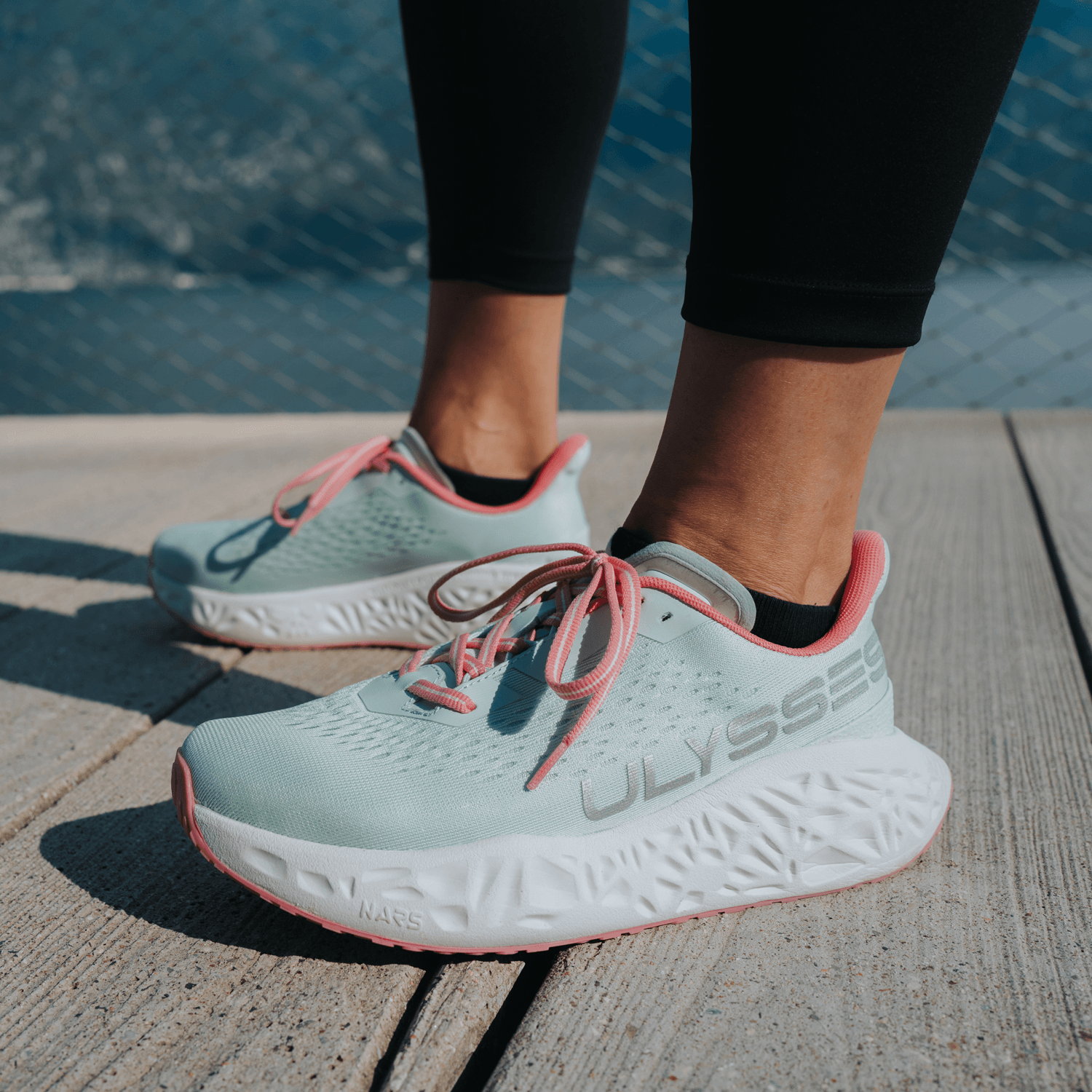 Close-up of light mint green Ulysses running shoes with pink laces and "NARS" branding on the midsole, worn on a wooden deck.