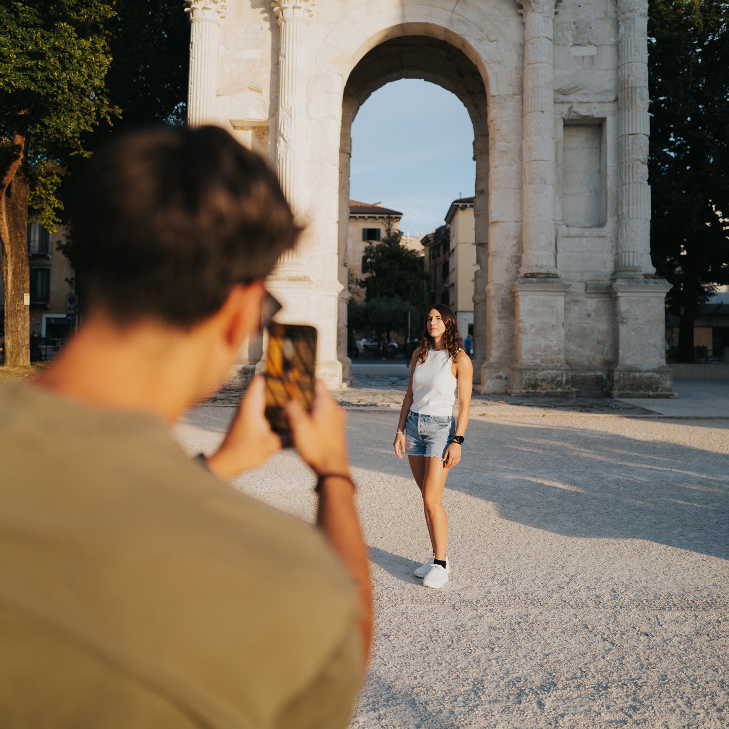 A lifestyle photo of a woman posing in front of a large stone archway, with a man in the foreground holding a smartphone to take the picture.