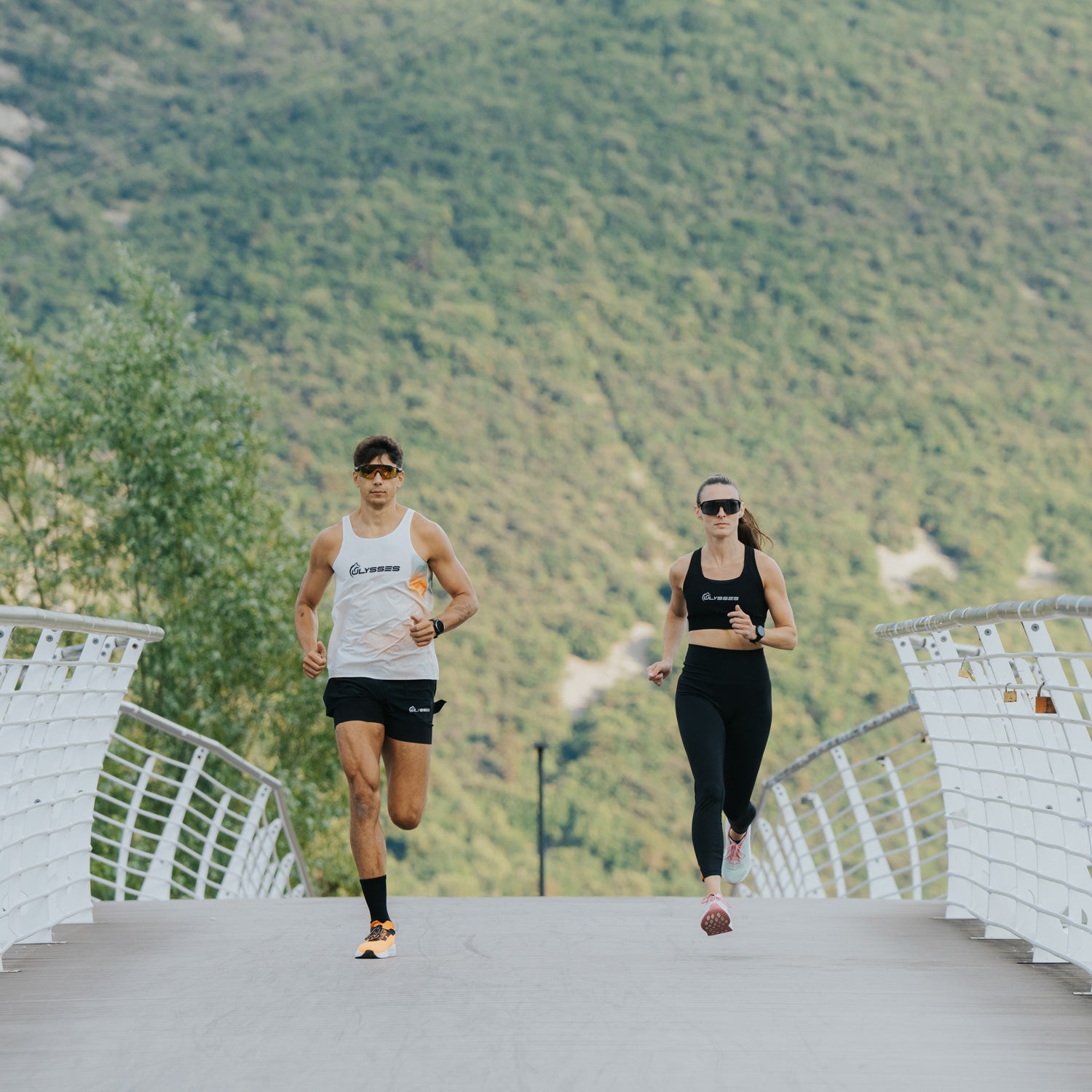 Two runners, a man and a woman, jogging on a modern white bridge with a lush green mountain backdrop.