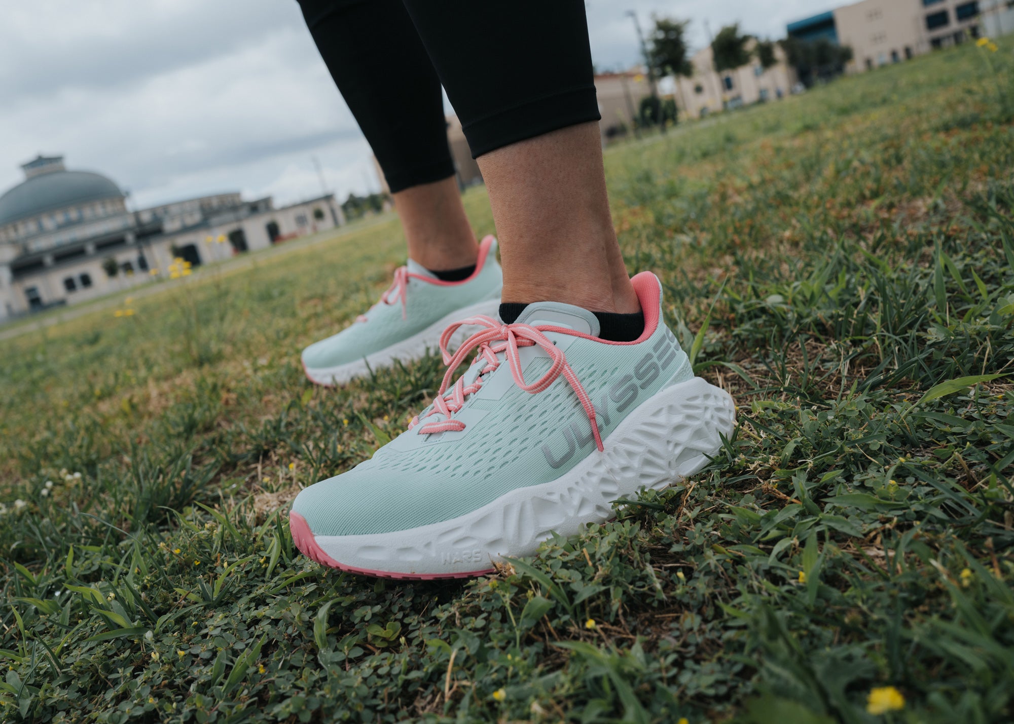 Low-angle close-up of a woman's feet wearing light mint and pink Ulysses running shoes, standing in a grassy field with buildings in the background.