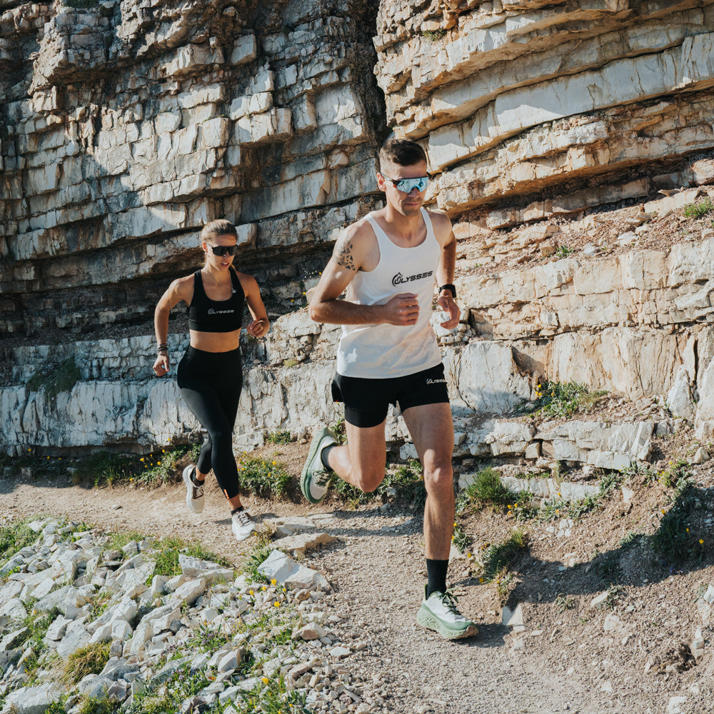 Two trail runners jogging on a rocky path with a large stone cliff behind them. 