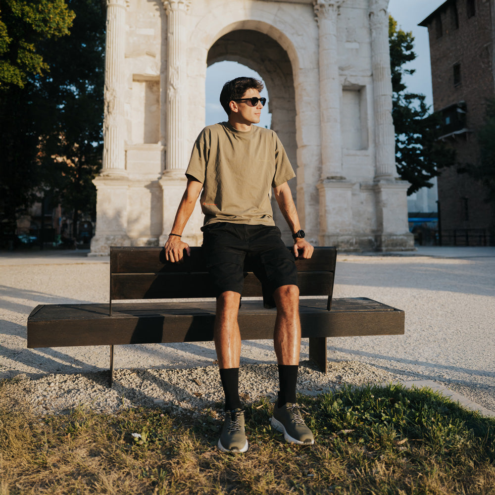 A man wearing a beige t-shirt, black shorts, and sunglasses sits on a dark wooden bench in front of a large stone archway.